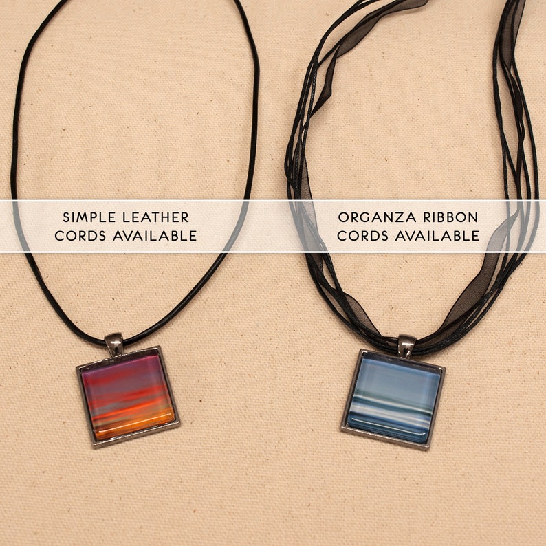 May include: Two necklaces with square glass pendants. The pendant on the left has a sunset-like design with orange, pink, and purple hues. The pendant on the right has a blue and white striped design. The necklace on the left is on a simple black leather cord. The necklace on the right is on a black organza ribbon cord. The text "SIMPLE LEATHER CORDS AVAILABLE" is above the left necklace. The text "ORGANZA RIBBON CORDS AVAILABLE" is above the right necklace.