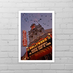 May include: A vintage-style movie theater marquee with neon lights advertising "HOLLYWOOD" and "Quentin Tarantino's The Hateful Eight in 70mm". The theater is decorated with ornate details and a clock tower. A flock of birds flies overhead against a twilight sky.