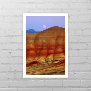May include: A full moon rises over a landscape of colorful, layered hills. The hills are a mix of red, orange, and yellow, with a blue sky in the background.