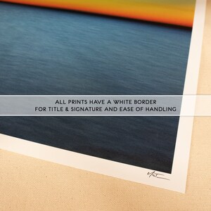 May include: A close-up of a print with a white border. The print features a blue and orange sunset over a body of water. The text "ALL PRINTS HAVE A WHITE BORDER FOR TITLE & SIGNATURE AND EASE OF HANDLING" is printed on the bottom of the print. The print is signed with the initials "V/ST".