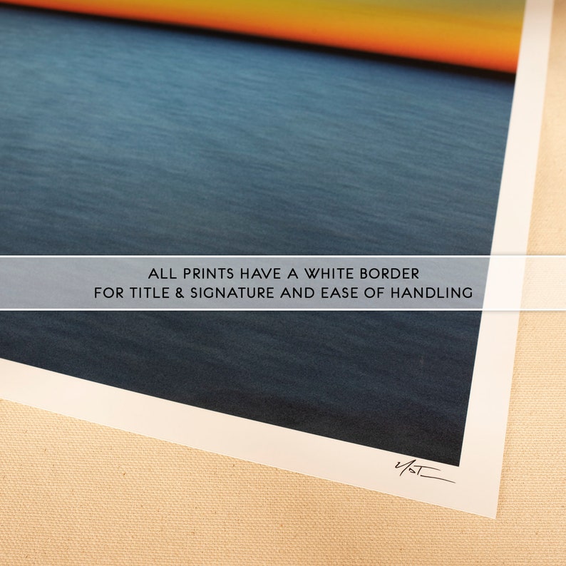 May include: A close-up of a print with a white border. The print features a blue and orange sunset over a body of water. The text "ALL PRINTS HAVE A WHITE BORDER FOR TITLE & SIGNATURE AND EASE OF HANDLING" is printed on the bottom of the print. The print is signed with the initials "V/ST".