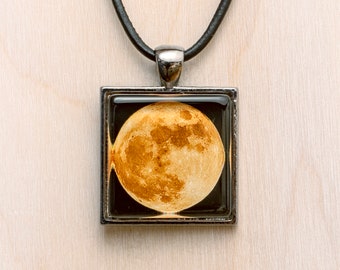 May include: A silver pendant necklace with a black leather cord. The pendant features a square frame with a clear resin insert showing a full moon in a dark sky.