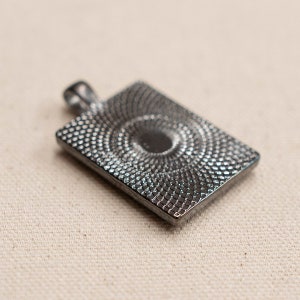May include: A silver-toned pendant with a square shape and a textured, circular design in the center.