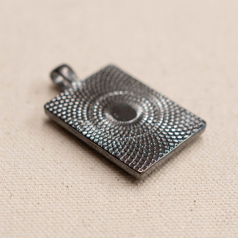May include: A silver-toned pendant with a square shape and a textured, circular design in the center.