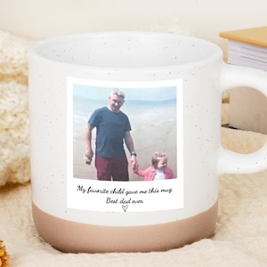 Può includere: Tazza in ceramica bianca con un design maculato e una base marrone. La tazza presenta una foto con il testo "My favorite child gave me this mug. Best dad ever" e un simbolo a cuore.