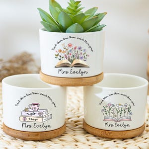 Puede incluir: Tres macetas de cerámica blanca con platillos de bambú. Cada maceta presenta un diseño floral y el texto "Teach them love them, watch them grow" y "Mrs. Evelyn". Una maceta contiene una planta suculenta.