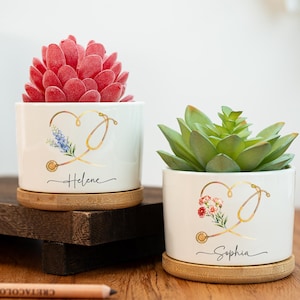 Puede incluir: Dos macetas de cerámica blanca con diseños dorados de estetoscopio y corazón, cada una con una suculenta. Una tiene una suculenta roja y el nombre "Helene", la otra una suculenta verde y el nombre "Sophia". Ambas están sobre platillos de bambú.