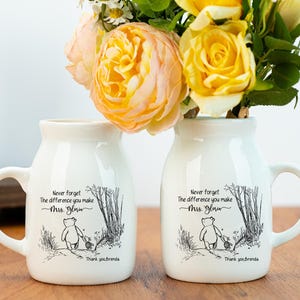 Puede incluir: Dos jarrones de cerámica blancos con asas, cada uno con una ilustración en blanco y negro de un oso y el texto "Never forget the difference you make Mrs. Gloria. Thank you Brenda." Los jarrones contienen flores amarillas y melocotón.