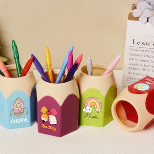 Puede incluir: Cuatro organizadores de escritorio con forma de lápiz en azul, morado, verde y beige, cada uno con un diseño y nombre únicos. Los organizadores contienen bolígrafos y lápices de colores. También hay un sacapuntas rojo y beige.