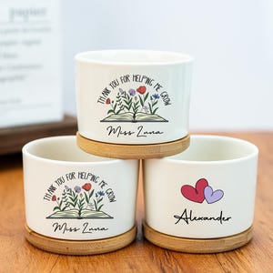 Può includere: Tre vasi da fiori in ceramica bianca con basi in legno. Il primo vaso ha un motivo floreale con il testo "Grazie per avermi aiutato a crescere Miss Luna". Il secondo vaso ha lo stesso motivo con il testo "Grazie per avermi aiutato a crescere Miss Luna". Il terzo vaso ha un motivo a cuore con il testo "Alexander".