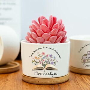 May include: A white ceramic planter with a pink succulent. The planter has a floral design with the text "Teach them love them watch them grow" and "Mrs. Evelyn". The planter sits on a wooden coaster.
