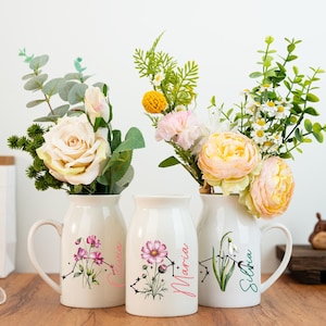 May include: Three cream ceramic vases with floral arrangements. Each vase has a handle and a floral design with a name. The flowers include roses, daisies, and other colourful blooms. The vases are on a wooden surface.