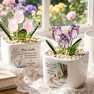 May include: Two white square ceramic planters filled with small stones and glass tulips. One planter has pink tulips and text reading Mrs. Smith, Thank you for being such an important part of my story, Love, Emily 2026. The other has purple tulips and text reading Mrs. Smith, Never forget the difference you make, Love, Emily 2026.
