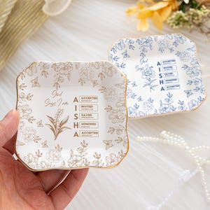 May include: Two square, floral-patterned trinket dishes. One is white with gold accents and a floral design, the other is white with blue floral patterns. Both have the text "God Says I Am" and the name "AISHA" with associated words.