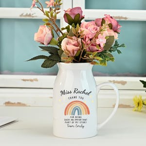 May include: A white ceramic jug with a handle, filled with pink and red flowers. The jug has text that reads "Miss Rachel, Thank You, For being such an important part of my story, From Emily" and a rainbow design.