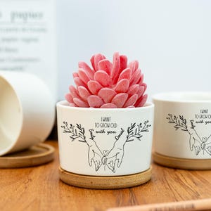 Könnte beinhalten: Weißer Keramik-Blumentopf mit einer rosa Sukkulente, auf einem Holzuntersetzer. Der Topf hat eine schwarze Illustration von Händen, die sich halten, mit dem Text "I want to grow old with you" und den Namen "Emily" und "Daniel".