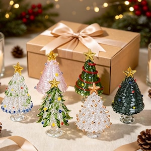 May include: A collection of six miniature glass Christmas trees in various colours including clear, pink, green, and white, each topped with a gold star and decorated with small colourful glass beads, arranged in front of a tan gift box with a ribbon.