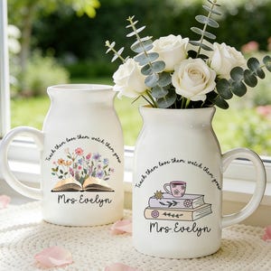 May include: Two white ceramic vases with handles, one holding white roses and eucalyptus. The vases are decorated with floral and book illustrations, the text "Teach them love them watch them grow" and "Mrs. Evelyn".