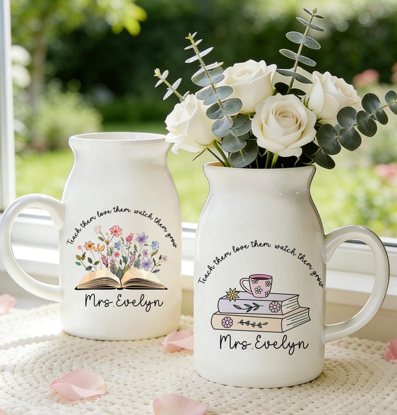 May include: Two white ceramic vases with handles, one holding white roses and eucalyptus. The vases are decorated with floral and book illustrations, the text "Teach them love them watch them grow" and "Mrs. Evelyn".