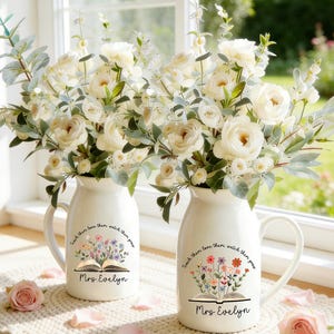 May include: Two white ceramic vases with handles, each holding a bouquet of white roses and greenery. The vases have a floral design with the text "Teach them love them watch them grow" and "Mrs. Evelyn".