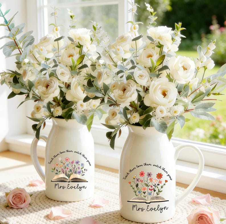 May include: Two white ceramic vases with handles, each holding a bouquet of white roses and greenery. The vases have a floral design with the text "Teach them love them watch them grow" and "Mrs. Evelyn".