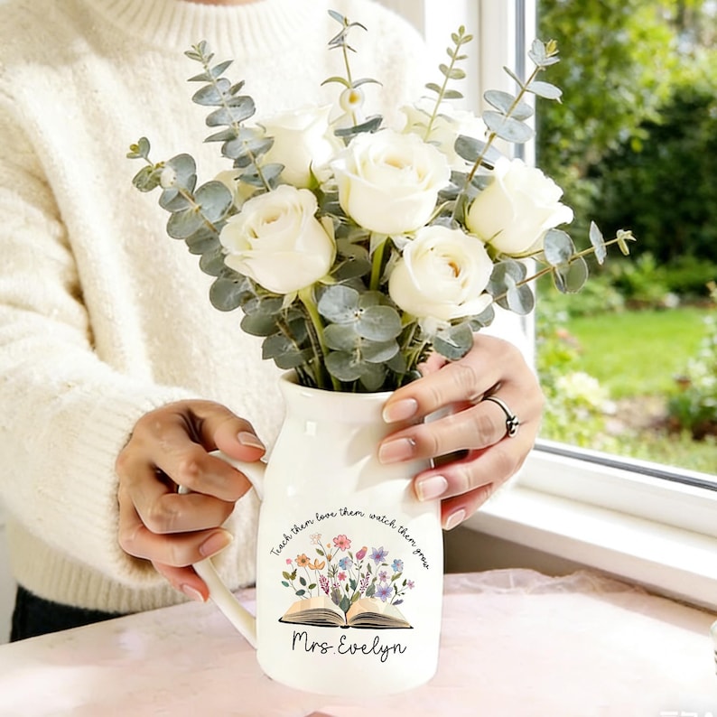 May include: A white ceramic pitcher with a handle, filled with white roses and eucalyptus. The pitcher is decorated with an open book illustration and the words "Teach them, love them, watch them grow" and "Mrs. Evelyn".