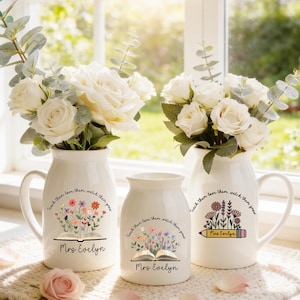 May include: Three white ceramic vases with floral arrangements. The vases have a handle and are decorated with the text "Teach them love them watch them grow" and "Mrs. Evelyn". One vase features an open book with flowers, another a pencil.