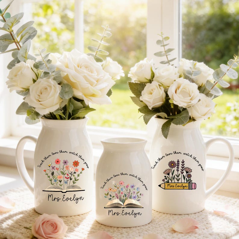 May include: Three white ceramic vases with floral arrangements. The vases have a handle and are decorated with the text "Teach them love them watch them grow" and "Mrs. Evelyn". One vase features an open book with flowers, another a pencil.