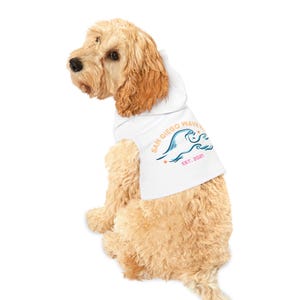 May include: A light-colored dog wearing a white hoodie. The hoodie features the text "SAN DIEGO WAVE" in a curved font, with a wave graphic in blue and pink, and the text "EST. 2021". The dog is sitting and looking to the left.