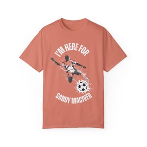 May include: A coral coloured t-shirt with a white graphic of a football player kicking a ball. The text "I'm here for Sandy Maciver" is printed on the shirt.
