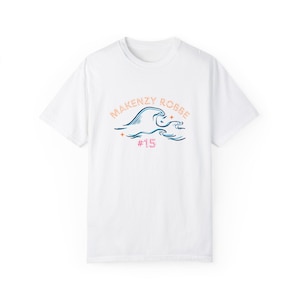 May include: White t-shirt with a blue and orange wave graphic and the text "MAKENZY ROBBE #15" printed on the front.