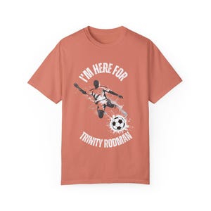 May include: A coral coloured t-shirt with a white graphic of a football player kicking a ball. The text "I'm here for Trinity Rodman" is printed on the shirt.