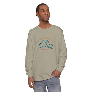 May include: A long-sleeved, light olive green shirt featuring a graphic of ocean waves with the text "SAN DIEGO WAVE FC" and "EST. 2021" in a blue and orange color scheme. The shirt is made of a soft material.