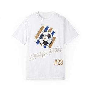 May include: White t-shirt with a blue and brown graphic of a soccer ball and the text "Jordan Bugs #23"