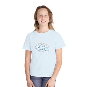 May include: A light blue t-shirt featuring the San Diego Wave FC logo. The logo includes a stylized wave design with the text "San Diego Wave FC" and "EST. 2021" in a circular arrangement. The shirt is made of a soft material.