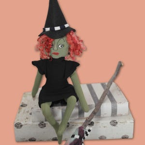 May include: A fabric doll with green skin, curly red hair, and a black witch hat featuring a black and white striped band. The doll wears a black dress and is accompanied by a small broomstick with a black and white striped handle accent.