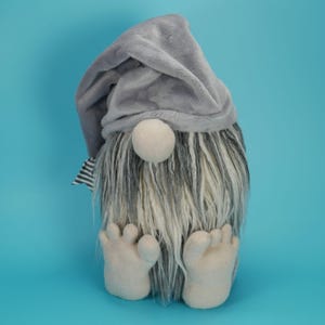 May include: A plush gnome figurine featuring a soft grey fabric hat, a round beige nose, and a long, shaggy beard with mixed grey and white fibers. The gnome has two beige fabric feet positioned at the front, and a small black and white striped tag.