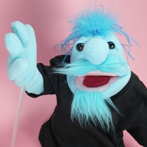 May include: A hand puppet with light blue fleece skin, a large nose, and messy blue hair. It features a blue moustache and goatee made of synthetic fibres, dark eyes, and an open mouth with a red tongue. The puppet wears a plain black shirt.