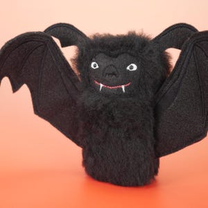 May include: A plush bat toy made of black fuzzy material with large, outstretched felt wings. The face features embroidered white eyes, a small nose, and a red stitched mouth with two white fangs protruding downwards from the upper lip.