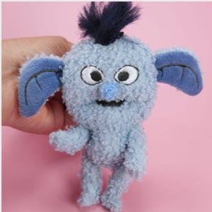 May include: A small, light blue plush creature with a fuzzy, curly texture. It features large white eyes with black pupils, a small blue nose, and a black stitched mouth. The creature has two large, smooth, dark blue ears and a tuft of dark blue hair.