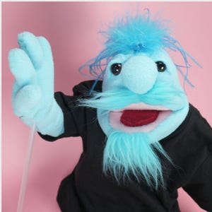 May include: A hand puppet with bright blue fleece skin, messy blue hair, a matching blue mustache and beard, and large black eyes. The puppet has an open mouth with a dark red tongue and is wearing a plain black long-sleeved shirt. A clear plastic rod is attached to one of the puppet's blue hands.