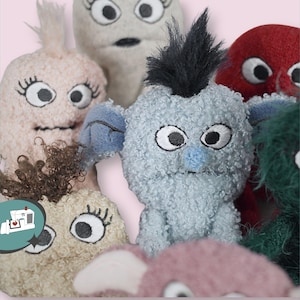 May include: A group of handmade plush monster toys in shades of pink, grey, blue, red, and green. Each monster has large, embroidered eyes and distinctive features. The toys are made from soft, textured fabric.
