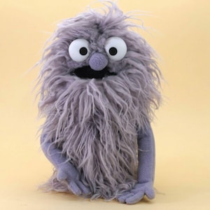 Könnte beinhalten: Eine Handpuppe mit langem, zotteligem, lavendelgrauem Kunstfell am Körper. Sie hat zwei große, runde weiße Augen mit schwarzen Pupillen, eine kleine lavendelgraue Stoffnase und einen breiten, offenen schwarzen Mund. Die Puppe hat zwei lavendelgraue Arme.
