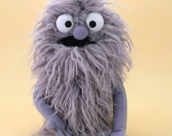 Könnte beinhalten: Eine Handpuppe mit langem, zotteligem, lavendelgrauem Kunstfell am Körper. Sie hat zwei große, runde weiße Augen mit schwarzen Pupillen, eine kleine lavendelgraue Stoffnase und einen breiten, offenen schwarzen Mund. Die Puppe hat zwei lavendelgraue Arme.