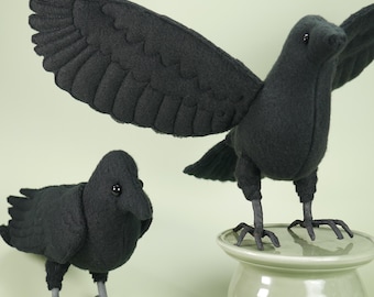 May include: Two black felt crow figures. One crow stands on a light green pedestal with its wings spread wide, while the other smaller crow stands on the surface. Both birds feature dark, glossy eyes and detailed stitched wing feathers.