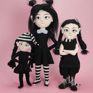 May include: Three plush dolls with black hair and pale skin, all wearing black outfits. The left doll wears a striped hat, the center doll has a white collar, and the right doll has braided hair with a striped bow and wears black lace-up boots.