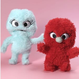 May include: Two plush toys with fuzzy textures. One is light blue with large embroidered eyes and long eyelashes. The other is bright red with large embroidered eyes, long eyelashes, and a small light blue circular nose centered on the face.