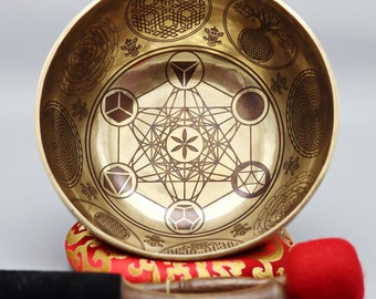 May include: A gold singing bowl with a geometric design, including a flower of life pattern, and two red and black mallets. The bowl is sitting on a red cushion with gold embroidery.
