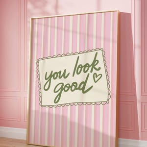 May include: A framed print with a pink and white striped background. The print features the phrase "you look good" in green script within a decorative border. The frame is gold-colored, and the print is leaning against a pink wall.