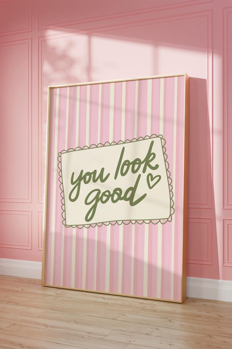 May include: A framed print with a pink and white striped background. The print features the phrase "you look good" in green script within a decorative border. The frame is gold-colored, and the print is leaning against a pink wall.